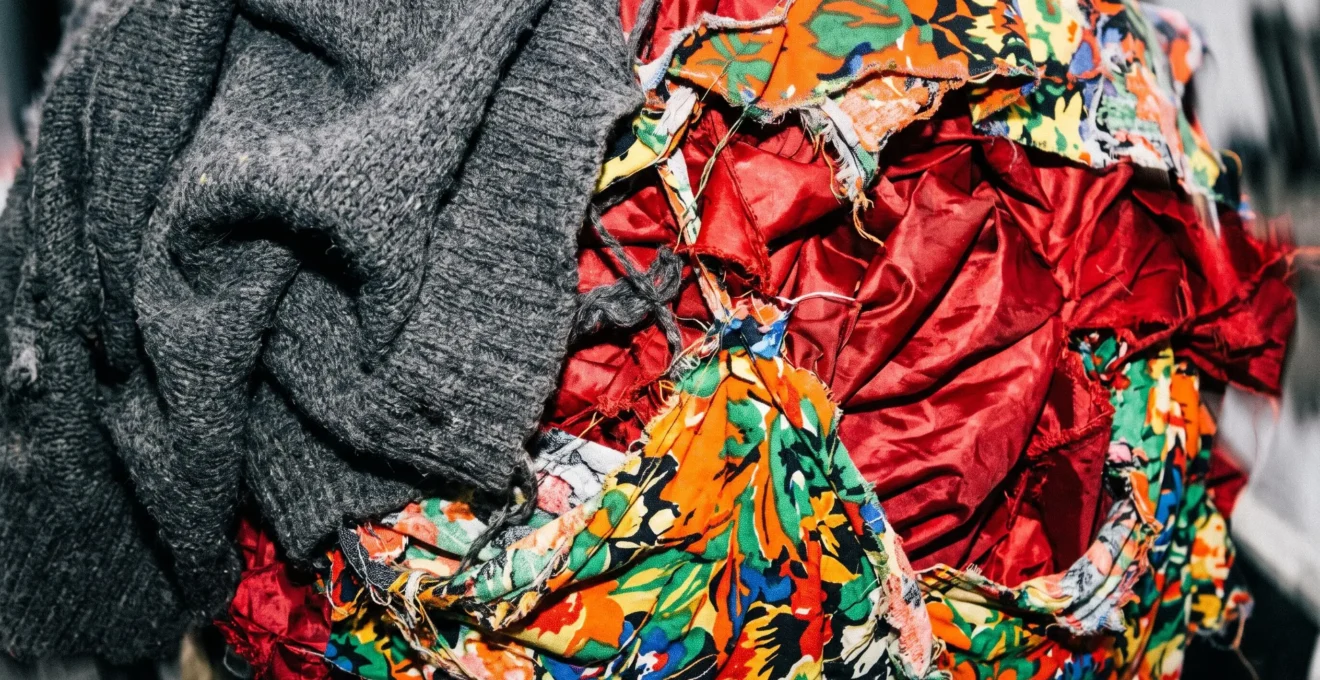 Composition abstraite chaotique de textiles en collision : maille laine grise, tissu synthétique rouge brillant et imprimé floral multicolore créant un ensemble visuellement discordant
